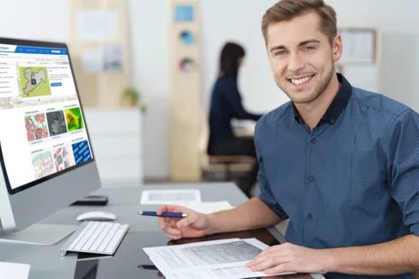 Ein junger lächelnder Mann sitzt vor einem Computer. Auf dem Computer ist eine Webseite des GeoSN geöffnet. Im Hintergrund befindet sich eine Frau.