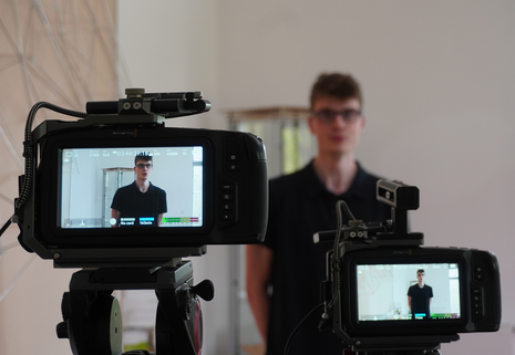 Das Bild zeigt zwei Kameras beim Videodreh für das Video der Laufbahnausbildung. Durch die Kamera ist ein junger Mann zu erkennen, welcher sich durch die Kameras Filmen lässt.