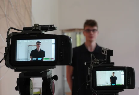 Das Bild zeigt zwei Kameras beim Videodreh für das Video der Laufbahnausbildung. Durch die Kamera ist ein junger Mann zu erkennen, welcher sich durch die Kameras Filmen lässt.